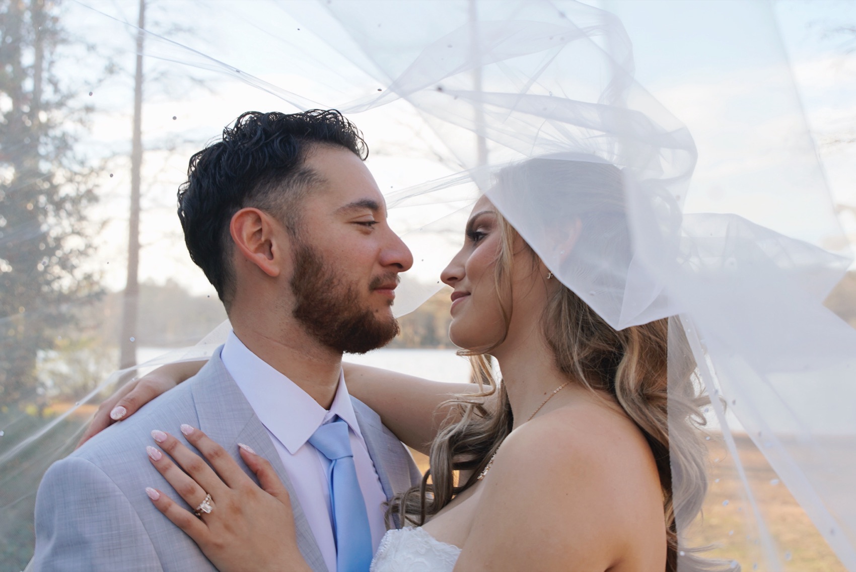 Bride and groom leaning in toward each other beneath her veil as it lifts in the wind by the lake at sunset